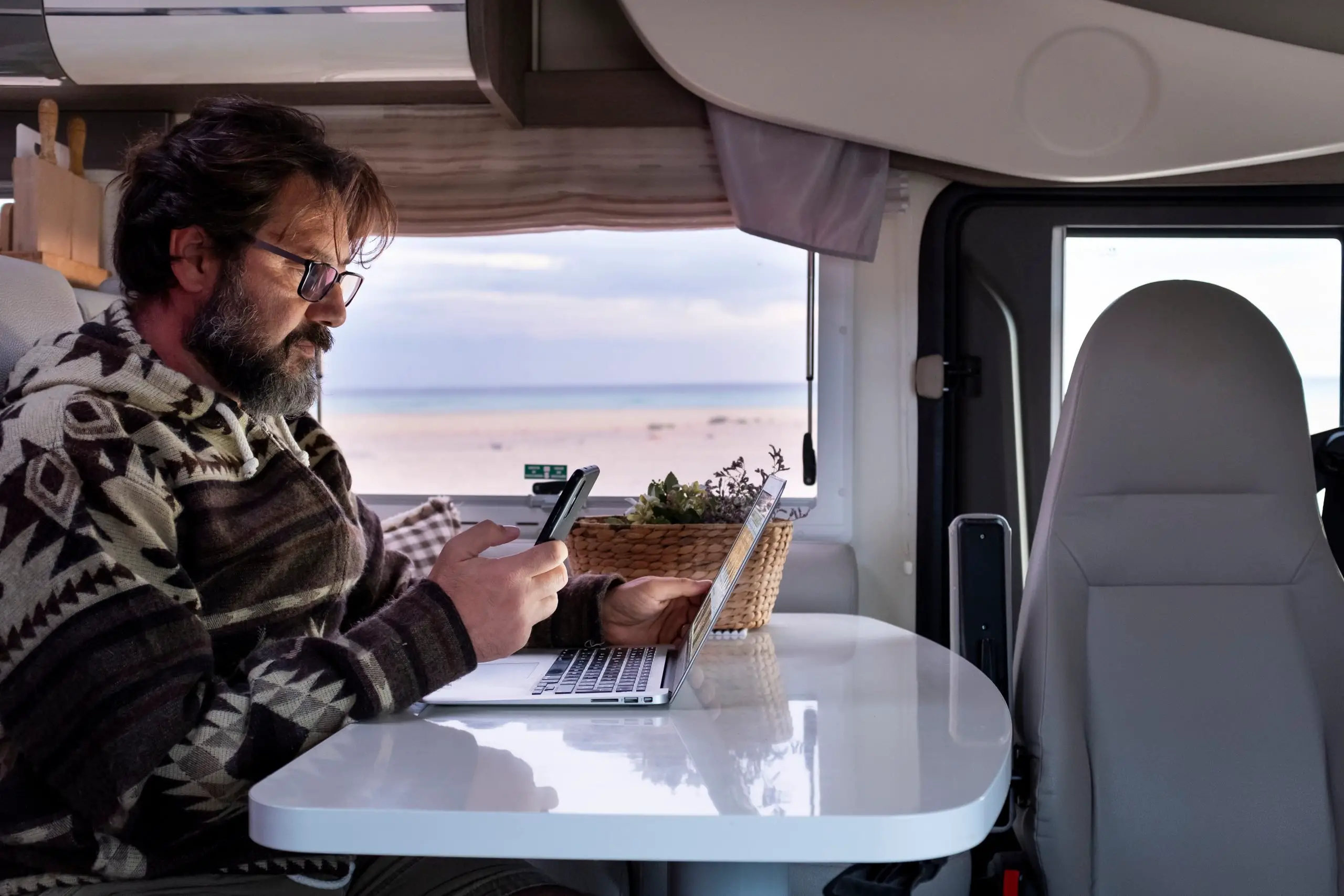 Man with cell phone in hand sitting at an RV table working on a laptop 