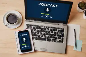 Laptop and cellphone displaying podcast beside a coffee cup on a desk.